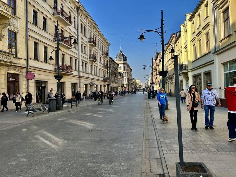 People walking along the historic Piotrkowska Street in Łódź, with classic architecture, street lamps, and a clear blue sky during the City Quest adventure.