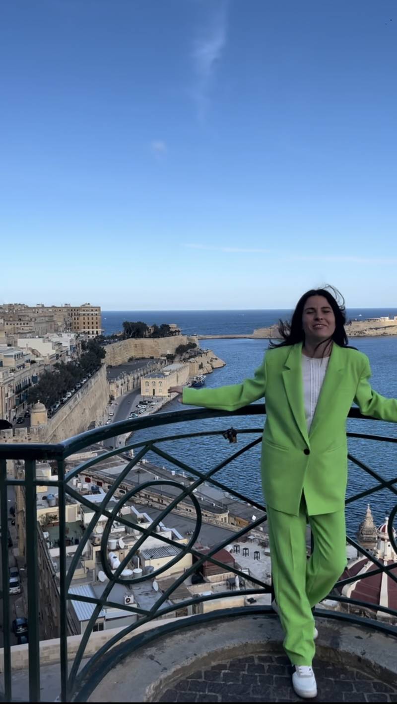Woman in bright green suit standing on balcony overlooking Valletta harbor and historic cityscape under clear blue sky