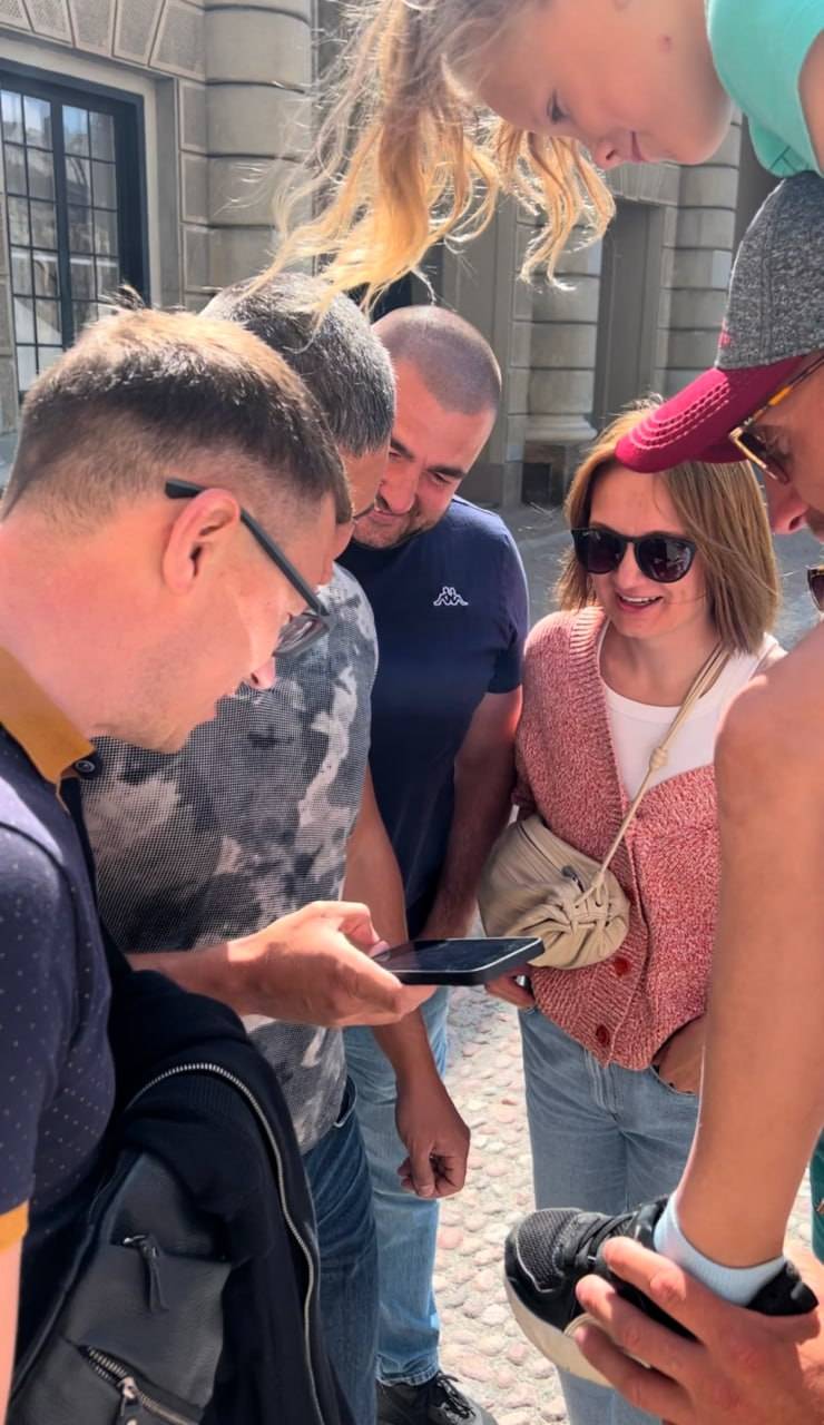 Gruppo di persone che esplora una città, guardando uno smartphone durante il City Quest a Lussemburgo, in un ambiente urbano soleggiato con edifici storici sullo sfondo.