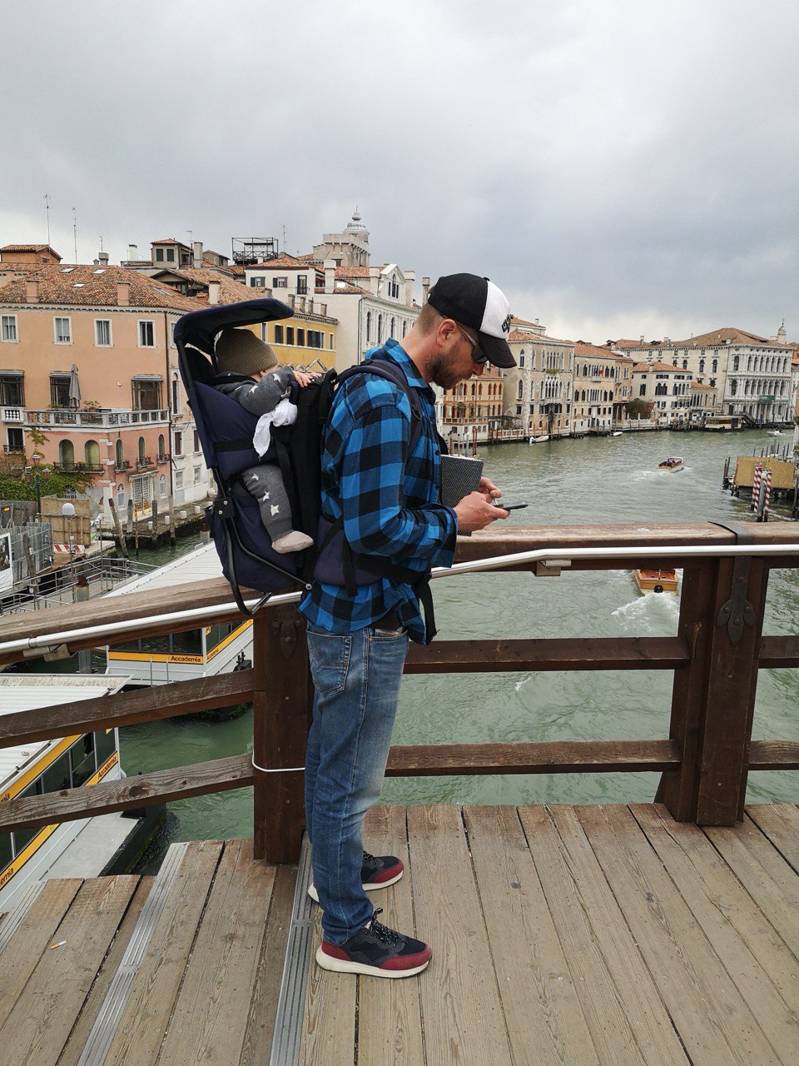 Man with a baby carrier using a smartphone on a wooden bridge overlooking Venice canals and historic buildings during the City Quest Venice adventure