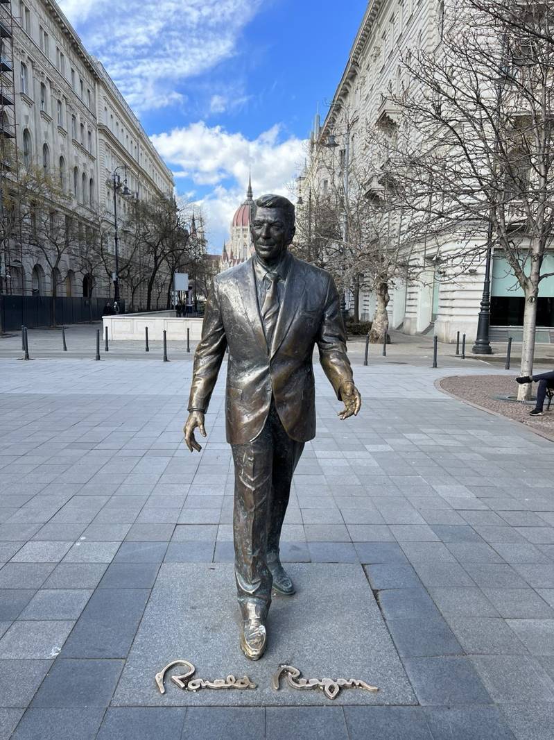 Bronze statue of Ronald Reagan walking on a paved square in Budapest with historic buildings and blue sky in the background