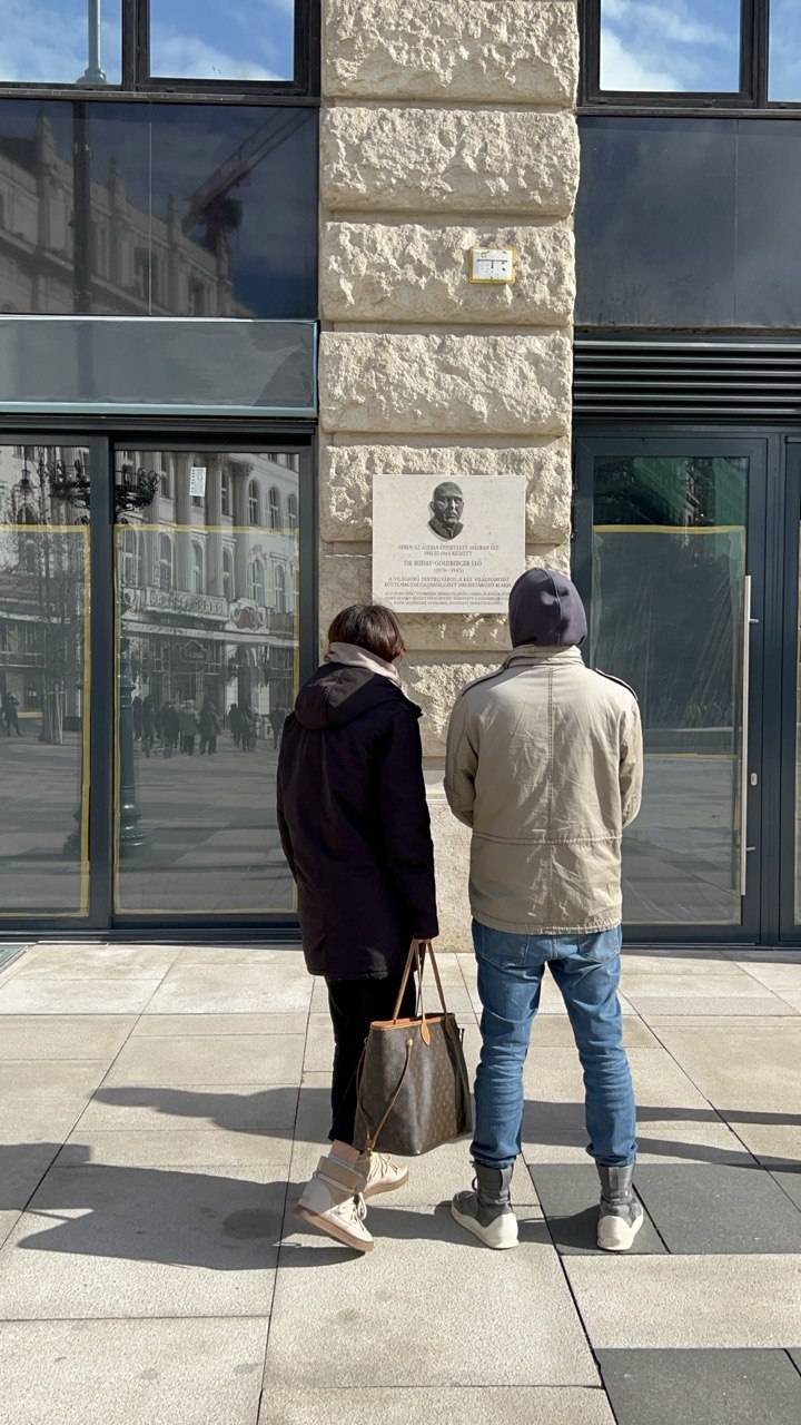 Two people standing and reading a commemorative plaque on a stone wall in Budapest during the City Quest, urban reflections visible in the windows nearby.