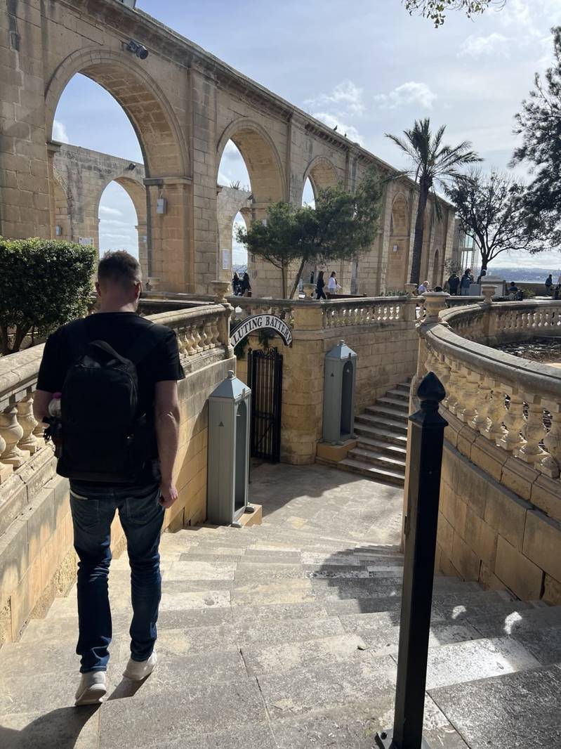 Man with backpack walking down stone stairs near historic arches and palm tree in sunny Valletta cityscape during city quest adventure