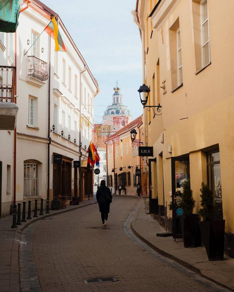 Narrow street in Vilnius with a person walking and colorful flags, historic buildings, and a distant view of Gediminas Tower under a clear sky.