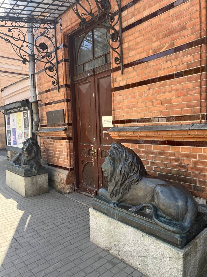 Вход в Дом Единства в Даугавпилсе с каменными скульптурами львов у двери на фоне кирпичной стены и кованого козырька над входом.