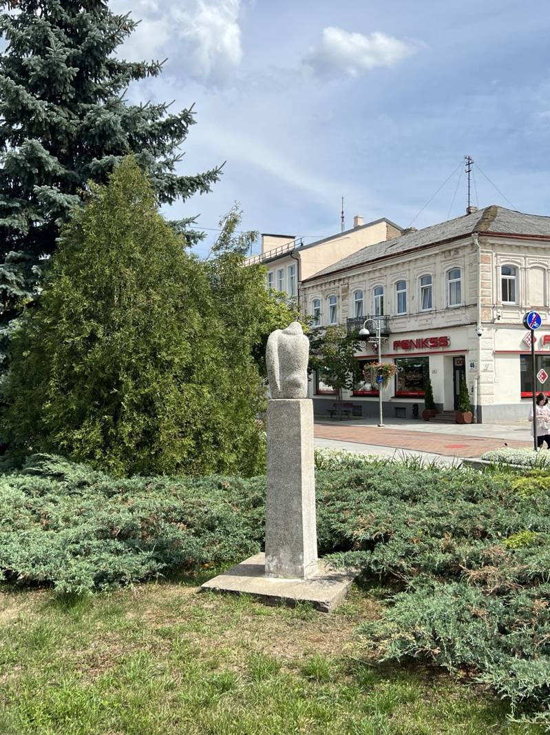 Скульптура на пьедестале в парке Даугавпилса на фоне зелёных кустов и старинных зданий с кафе, частично затенённая деревьями под голубым небом с облаками.